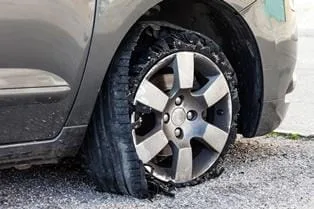 A car with a flat tire is parked on gravel, its sidewall damaged and torn from an apparent tire blowout.