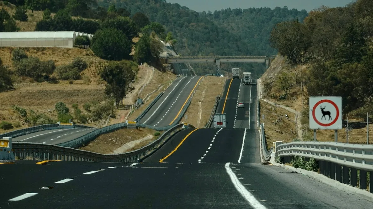 A highway with multiple lanes passes through hilly terrain. There are vehicles on the road and a deer-car accident lawyer warning sign visible on the right side, alerting drivers to potential wildlife crossings.