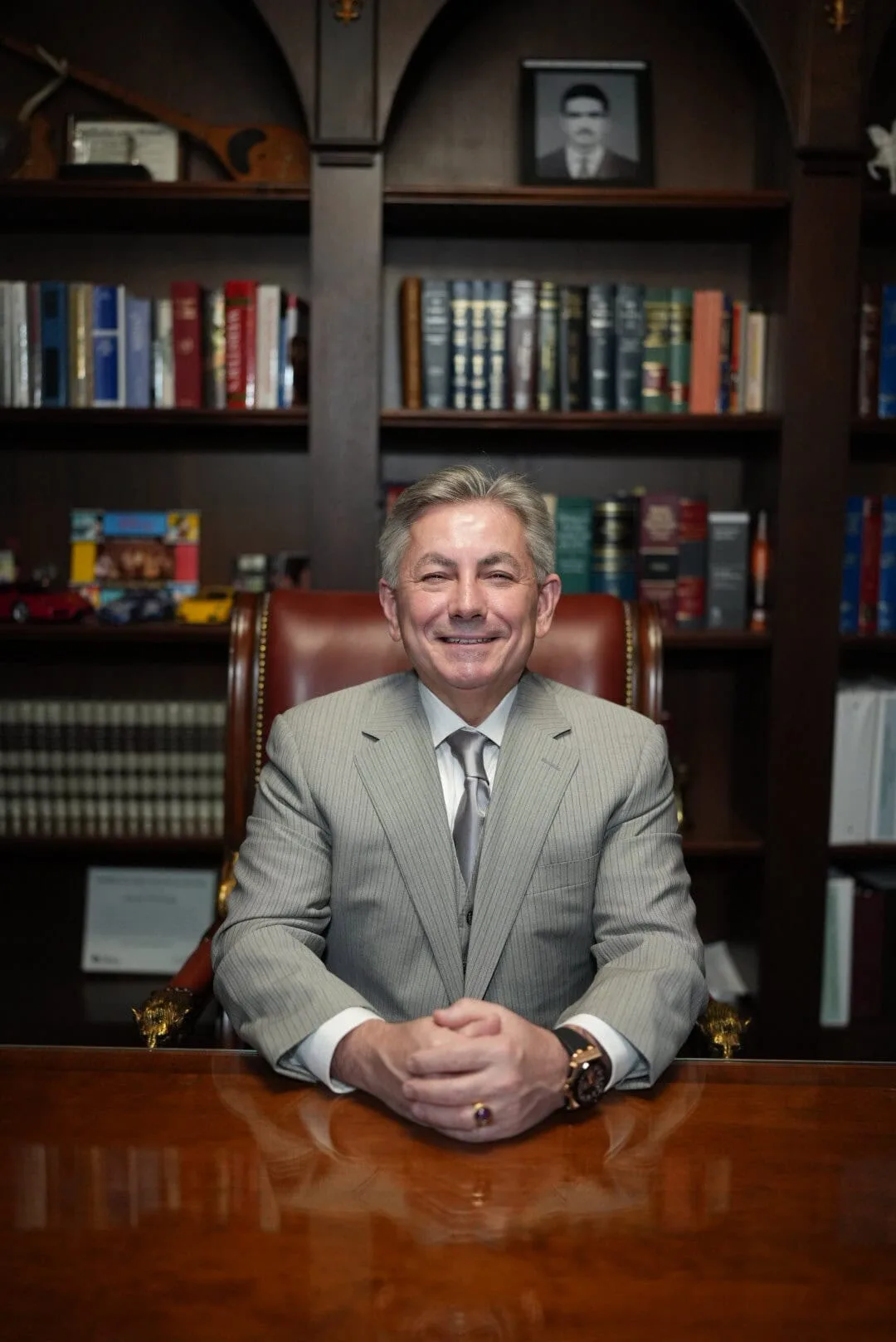 Joseph Dedvukaj seated at his desk in the firm's office, smiling, with bookshelves and a framed photo in the background