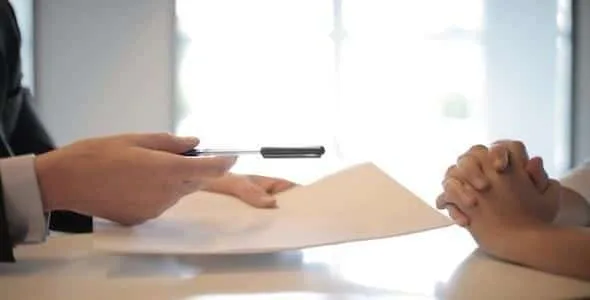 A person in a suit is handing over a settlement offer document and pen to another, whose hands are clasped on the table.