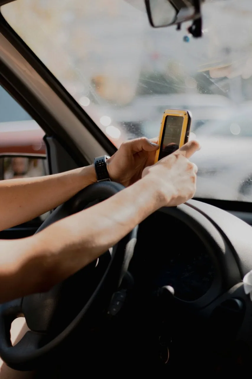 A person in the driver’s seat holds a smartphone above the steering wheel inside a car, sunlight streaming through the windshield—a scene often addressed by a Distracted Driving Lawyer.