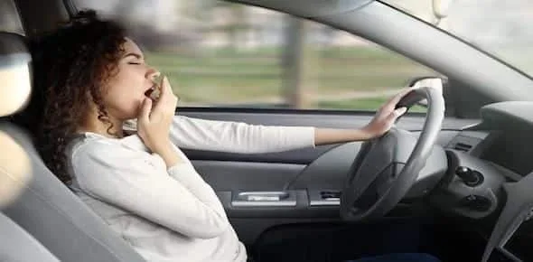 A person in the driver's seat yawns with one hand on the steering wheel, a clear sign of drowsy driving.