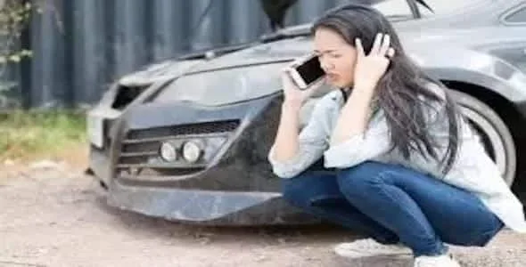 A woman is squatting beside a damaged car, talking on the phone and appearing distressed after a Michigan car accident.