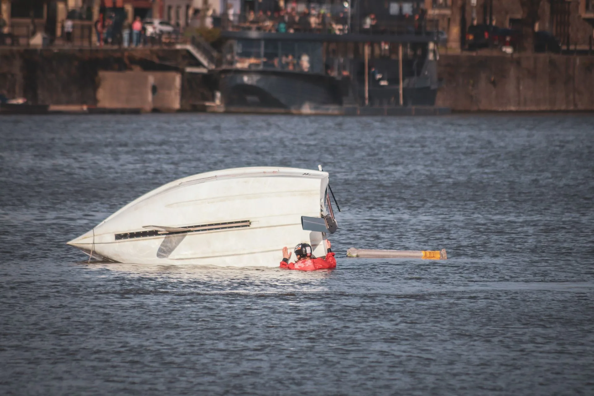 Capsized white boat with a person in a red survival suit in the water beside it