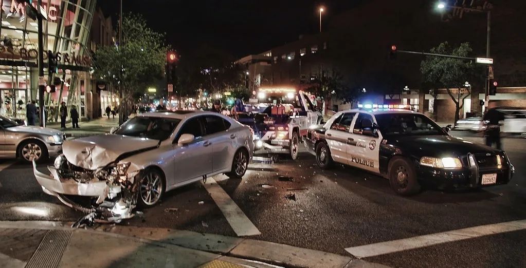 In a late-night incident at a city intersection, two cars, including a police vehicle, are damaged in what appears to be a drunk driving accident. The scene is illuminated by the flashing lights of a tow truck and police vehicles in the background.