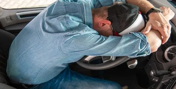 Person in denim shirt and cap rests head on car steering wheel, arms folded over head, a clear sign of driver fatigue.