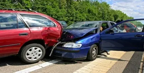 A blue car rear-ends a red car on a tree-lined road, causing visible damage from the car accident.