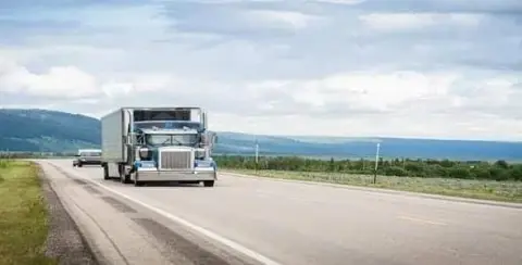 A blue semi-truck cruises along an open highway, framed by majestic mountains and cloudy skies. If you're dealing with truck driver negligence, consider a free consultation to navigate your next steps with ease.