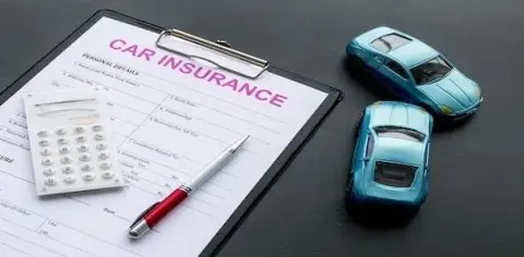 A car insurance form, accompanied by a pen and calculator, sits neatly on a gray surface alongside two blue toy cars. Ensure you're well-informed about your coverage to avoid mistakes.
