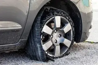 A car with a flat tire is parked on gravel, its sidewall damaged and torn from an apparent tire blowout.
