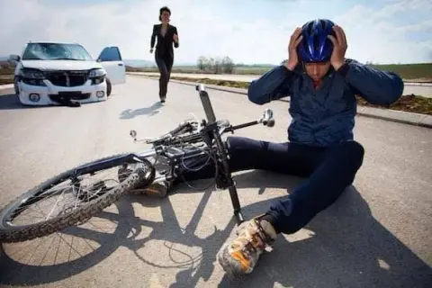 A cyclist sits on the road clutching their helmet by a fallen bicycle, indicating a bike accident, with a car and a person rushing towards them in the background.