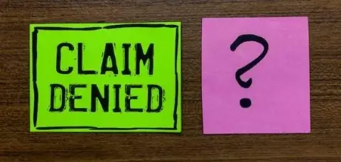A green note reads "CLAIM DENIED" next to a pink note with a question mark on a wooden surface, echoing the frustration of an auto accident claim in Michigan involving an aggravated pre-existing medical condition.
