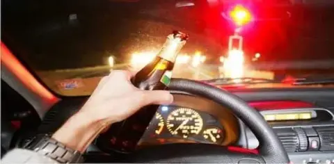 A hand grips a beer bottle while driving at night, hinting at a reckless choice made by a drunk driver. The blurred view of the speedometer and the ominous red traffic light ahead underscore the looming danger and potential for an injury claim.