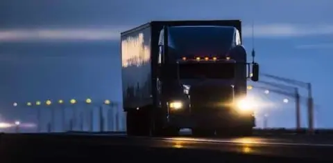 A large truck, piloted by a vigilant long haul truck driver with headlights on, travels down a highway at dusk, blurred streetlights painting the backdrop.