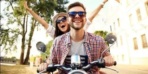 A man and woman ride a motorcycle, both smiling and wearing sunglasses. The woman raises her arms joyfully, embracing the thrill of the ride. Trees and buildings blur in the background, as they enjoy a carefree day without a hint of road rash or concern for motorcycle accidents.