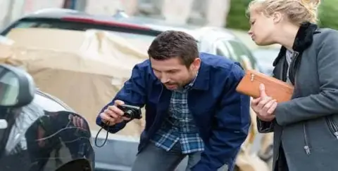 A man crouches to photograph the car at a crash scene, while a woman with a purse looks on. Cars with protective covers are in the background.