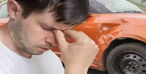 A man in a white shirt holds his face in frustration after a minor accident. Behind him, an orange car, damaged on the side, awaits an insurance claim.