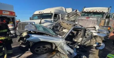 A mangled silver car is crushed between two large semi-trucks on a highway. Emergency responders are visible at the scene, highlighting the severity of the personal injury involved in this car rear-end truck accident.