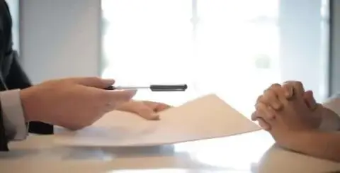 A person in a suit is handing over a settlement offer document and pen to another, whose hands are clasped on the table.