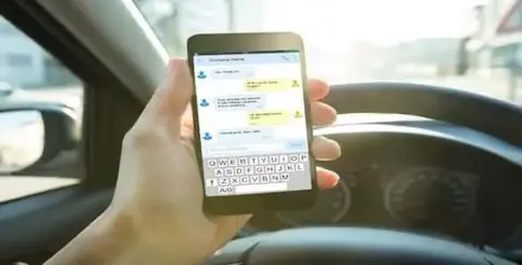 A person seated in a car is using a smartphone to display a chat conversation, with the steering wheel clearly visible, highlighting the potential for distracted driving accidents.