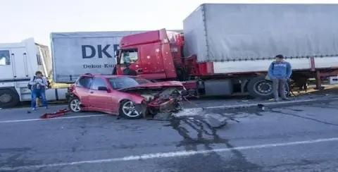 A red car with a severely damaged front sits in front of a large truck on the road, capturing the concern of two onlookers. As they assess the situation, thoughts might turn to Michigan Highway Accident Attorneys for expert guidance.