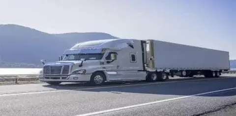 A silver semi-truck, one of the common types of big trucks, sits parked on a highway, with a long trailer and majestic mountains beneath a clear sky in the background.
