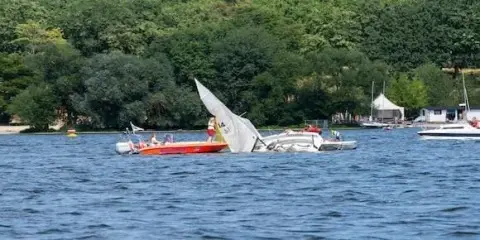 A small sailboat, capsized, is partially submerged in a lake as a motorboat with people nearby observes. Trees and a building are visible in the background, highlighting the importance of boating safety in such serene settings.