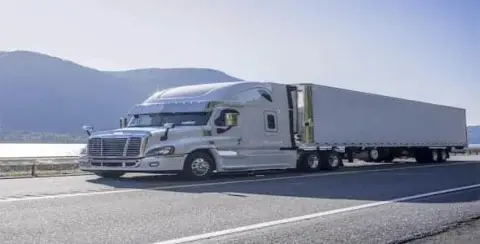 A white semi-truck with a long trailer navigates the highway, framed by mountains and a shimmering body of water in the background, evoking thoughts of trucking accident claims.