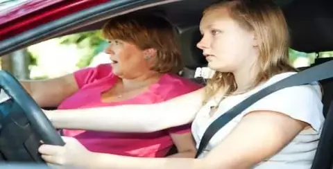 A woman watches a teen driving a car, both looking forward, with the young driver focused on the road. This careful observation aligns with graduated student driver licensing laws, ensuring teens gain confidence behind the wheel under parental guidance.