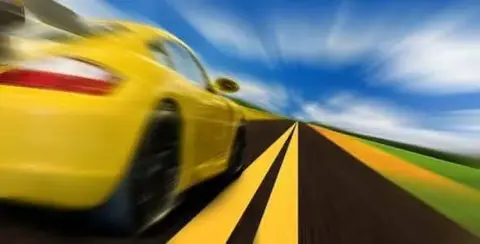 A yellow sports car, pushing the limits of excessive speed, zooms down the road with motion blur against a backdrop of blue sky and clouds.