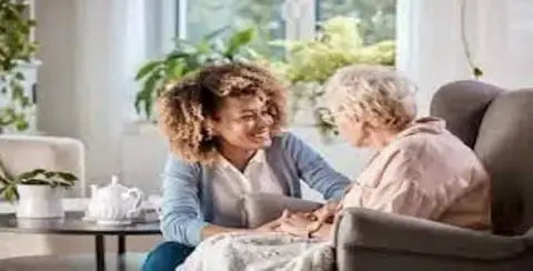 A young woman and an elderly woman sit together in a cozy living room, smiling and holding hands, reminiscent of the compassion seen in Michigan Attendant Care services.