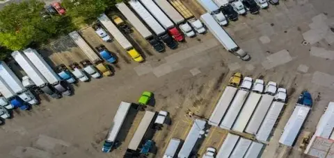 Aerial view of a trucking company lot with numerous parked semi-trucks and trailers in neat rows. Some trees and various colors of truck cabs are visible, showcasing the diverse fleet managed by experienced truck drivers.