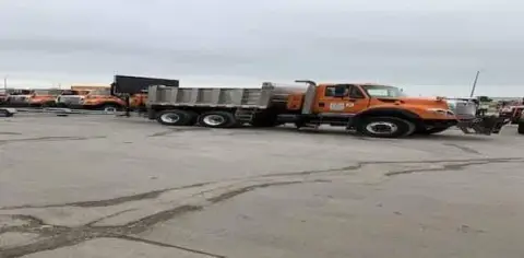 An orange utility truck, possibly a salt truck, is parked in a lot with other similar vehicles under an overcast sky.