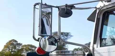 Close-up of a truck's side mirrors and a red signal light, highlighting potential blind spots, with trees and a clear blue sky in the background.