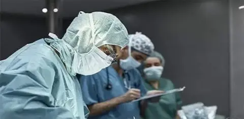 Doctors in scrubs and masks are meticulously performing surgery in an operating room, a procedure necessitated by a severe car accident. Two of them are diligently taking notes, ensuring every detail is accounted for during the operation.