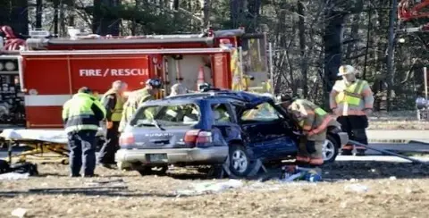 Emergency responders are at the scene of an angle collision car accident involving a blue vehicle and a fire rescue truck in a wooded area.