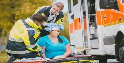 Paramedics assist a seated woman near an ambulance, tending to her with medical equipment after a car accident.