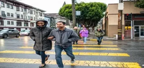 People walk across a crosswalk on a rainy day, with two individuals in front arm-in-arm while others follow behind. Buildings and trees line the street, offering a typical city scene where staying aware of pedestrian accident dos & don'ts is crucial for safety.