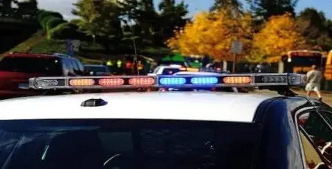 Police car with flashing lights at a roadside scene; other vehicles and people visible in the background on a sunny day with autumn trees, as authorities conduct a truck accident investigation.