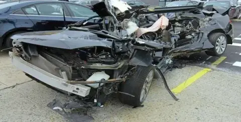 Severely damaged car with a crumpled front and exposed interior, likely the result of a harsh accident, parked on a street near other vehicles.