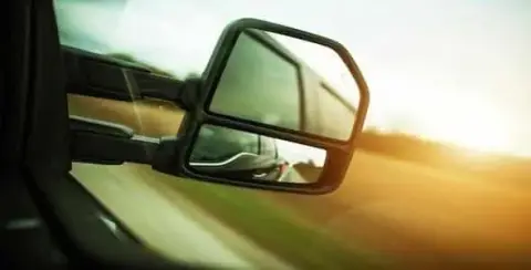 The side mirror of a vehicle reflects a serene road and countryside landscape, with sunlight in the background—a reminder of how crucial it is for Michigan Blind Spot Truck Accident Lawyers to address unseen dangers.