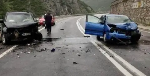Two cars with significant front-end damage rest on a two-lane road after a collision, debris scattered around. In the aftermath of what appears to be a Michigan car accident, two people are walking away from the scene.