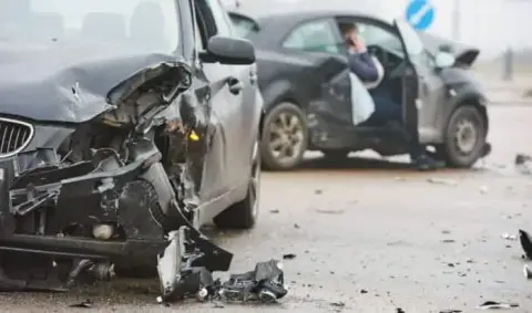 Two damaged cars on a road following a collision, perfectly illustrating the chaos often encountered when reporting car accidents. The front of the car in the foreground is significantly crumpled, debris is scattered around, and a person is sitting in the other car.