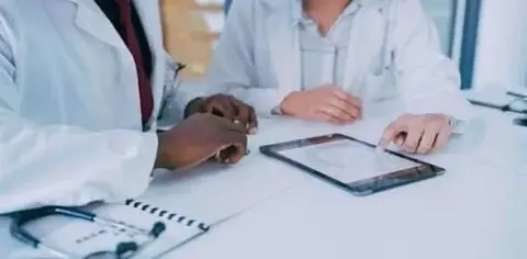 Two doctors in white coats discuss the importance of medical care information on a tablet at a table with a notepad and stethoscope nearby.