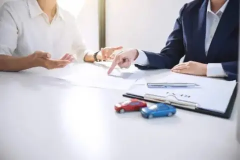 Two people discuss documents at a table, one pointing at papers while the other gestures animatedly. Nearby are toy cars and a clipboard with a pen, perhaps suggesting they’re planning social media posts about a car accident or vehicle safety tips.