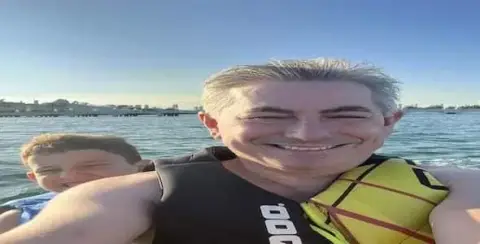 Two people wearing life jackets smile while enjoying a Michigan boating adventure, with water and distant buildings enhancing the backdrop.