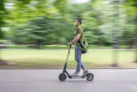 Wearing a green mask and shirt, the person skillfully navigates their Detroit electric scooter swiftly through the park, mindful of sidewalk safety.