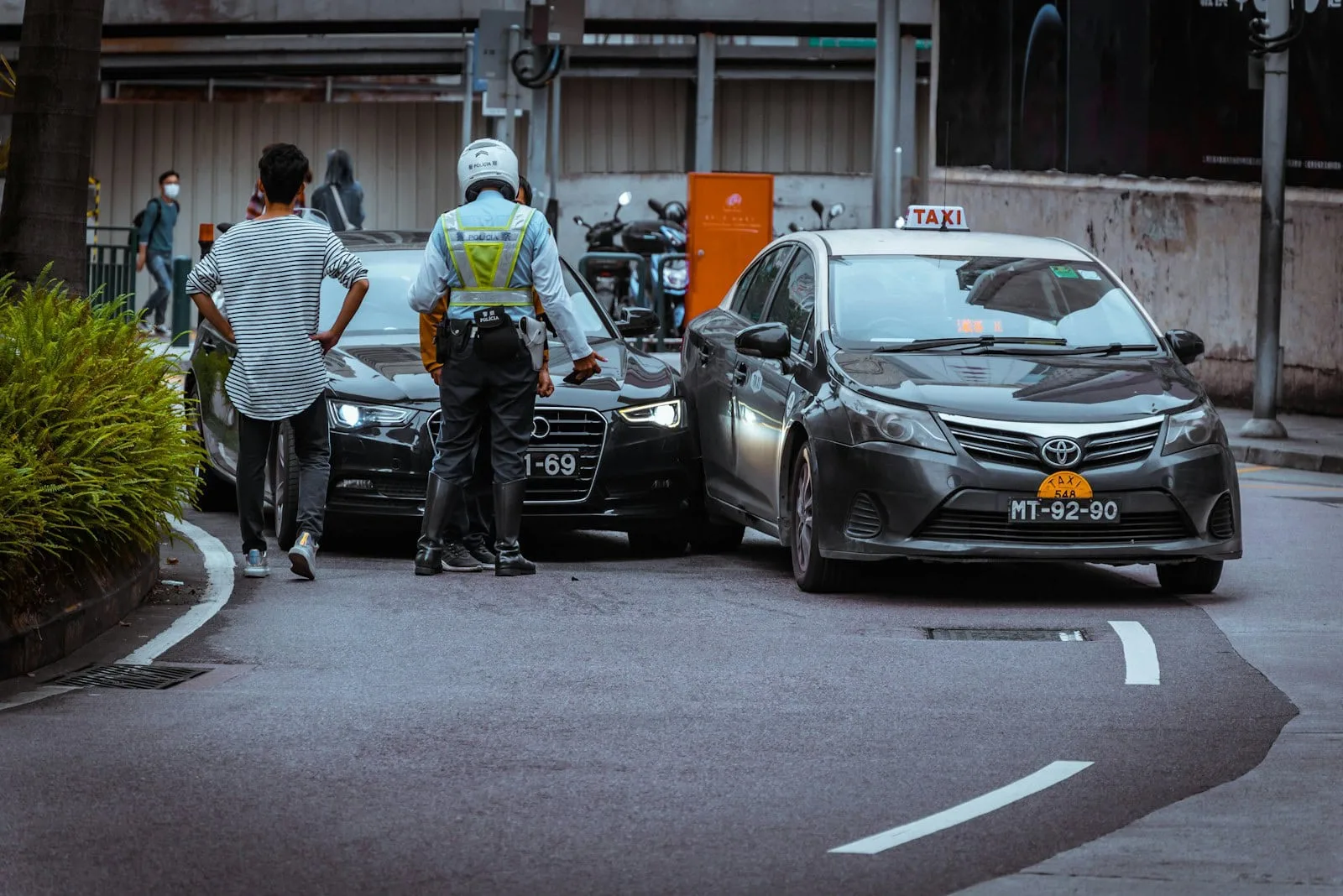 Two cars, a Mercedes and a Toyota, have been involved in a minor rear-end collision on the street. A person and a traffic officer are standing nearby.