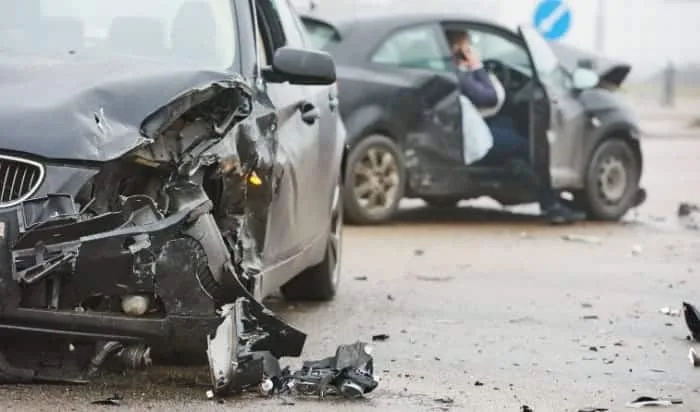 Two damaged cars on a road following a collision, perfectly illustrating the chaos often encountered when reporting car accidents. The front of the car in the foreground is significantly crumpled, debris is scattered around, and a person is sitting in the other car.