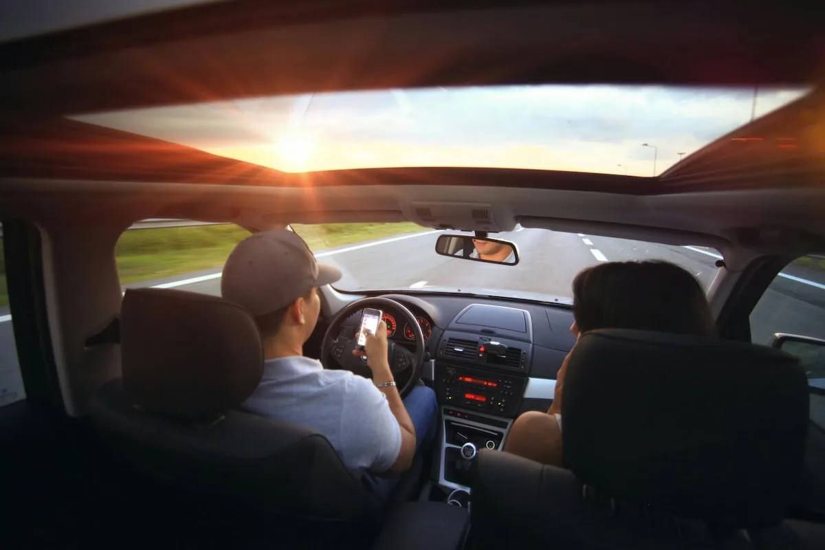 Two people sit in the front seats of a car driving on an empty road at sunset; the driver looks at a phone while steering, highlighting a scene where consulting a distracted driving lawyer might be necessary.